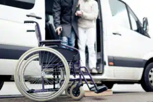 foto ravvicinata di una carrozzina per disabili con dietro il pulmino per il trasporto disabili e un operatore che aiuta una persona anziana a scendere