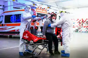 Due operatori sanitari con tutta integrale, mascherina e occhiali protettivi, visitano un paziente durante il covid seduto su una barella con alle spalle l'ambulanza. 